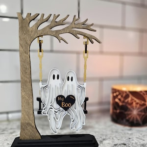 May include: A wooden Halloween decoration featuring two white ghosts on a swing, with the words "My Boo" on a black heart. The swing hangs from a wooden tree, all set on a black base.