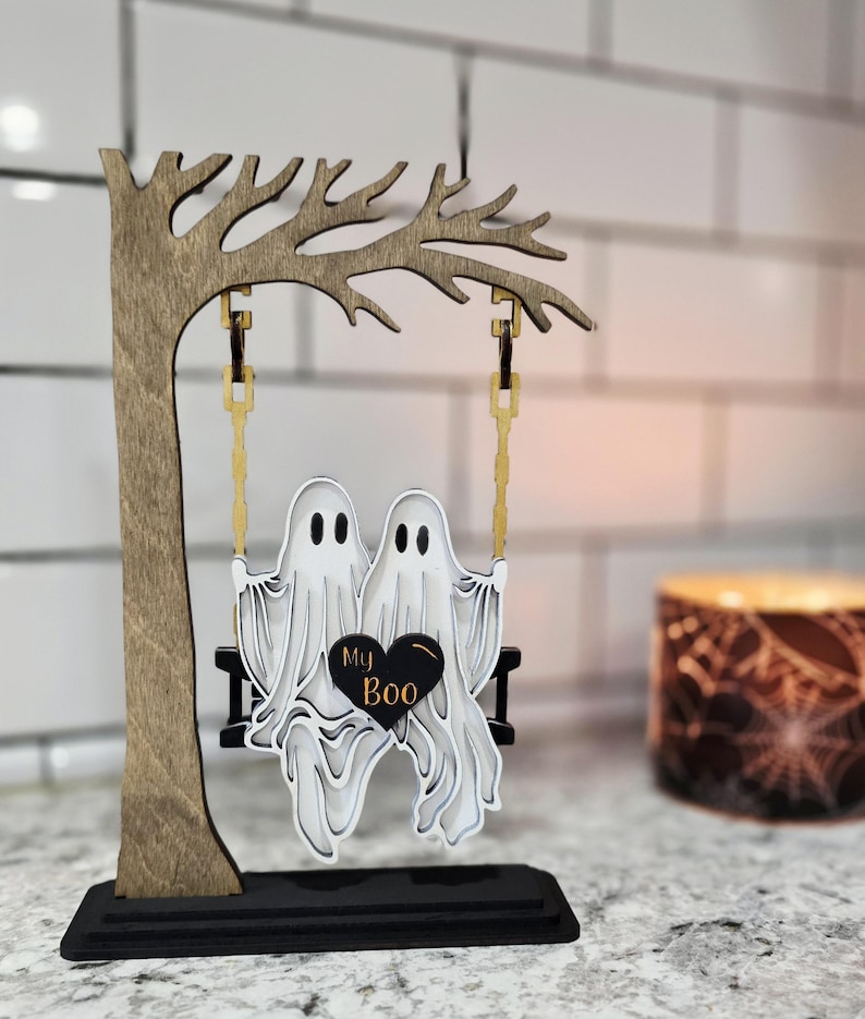May include: A wooden Halloween decoration featuring two white ghosts on a swing, with the words "My Boo" on a black heart. The swing hangs from a wooden tree, all set on a black base.