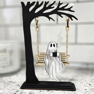 May include: A wooden Halloween decoration featuring a ghost on a swing. The ghost is white and holding a black mug, with steam rising. The swing is attached to a black tree, all on a black base.