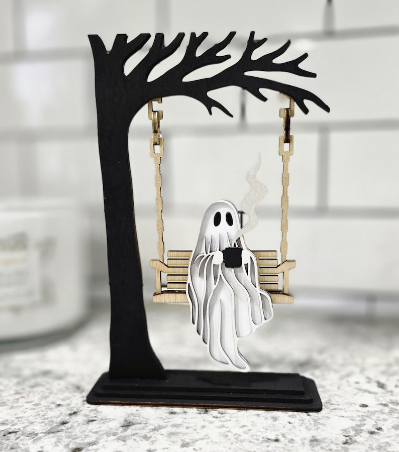 May include: A wooden Halloween decoration featuring a ghost on a swing. The ghost is white and holding a black mug, with steam rising. The swing is attached to a black tree, all on a black base.