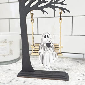 May include: A Halloween-themed wooden decoration featuring a black tree with a swing. A white ghost figure is seated on the swing, holding a black mug with steam rising. The base is rectangular.