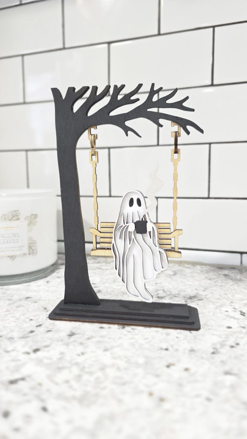May include: A Halloween-themed wooden decoration featuring a black tree with a swing. A white ghost figure is seated on the swing, holding a black mug with steam rising. The base is rectangular.