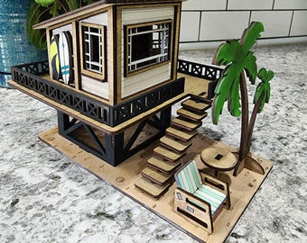 May include: A wooden miniature of a lifeguard station with a palm tree, chair, and table. The station has a balcony with two surfboards. The structure is painted in shades of brown, white, and black. The base is a light tan color.