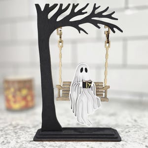 May include: A Halloween-themed wooden decoration featuring a black tree with a swing. A white ghost figure is seated on the swing, reading a book. The ghost and swing are made of layered wood, creating a 3D effect. The base is rectangular.