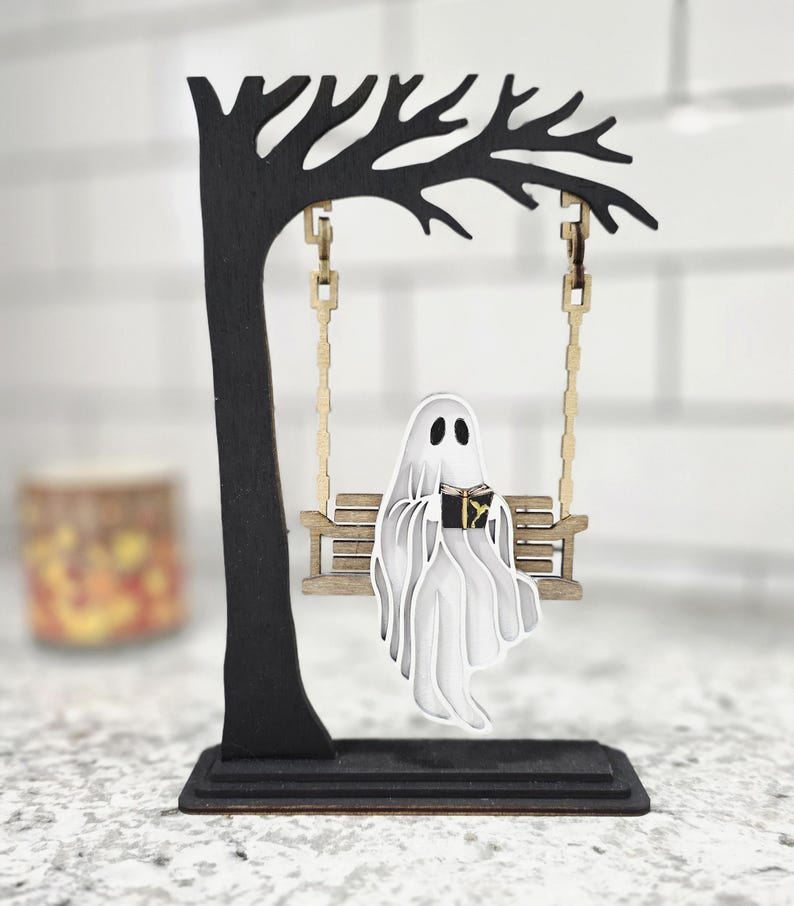 May include: A Halloween-themed wooden decoration featuring a black tree with a swing. A white ghost figure is seated on the swing, reading a book. The ghost and swing are made of layered wood, creating a 3D effect. The base is rectangular.
