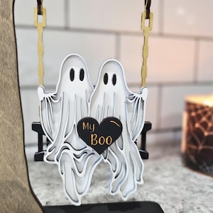 May include: A wooden Halloween decoration featuring two white ghost figures on a swing, with a black heart that says "My Boo." The swing hangs from a wooden tree. The base is black.