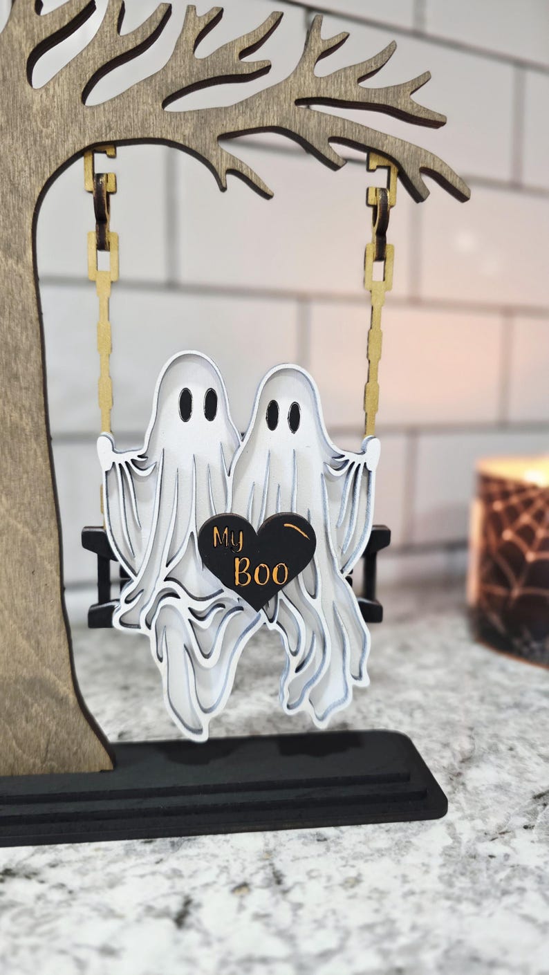 May include: A wooden Halloween decoration featuring two white ghost figures on a swing, with a black heart that says "My Boo." The swing hangs from a wooden tree. The base is black.