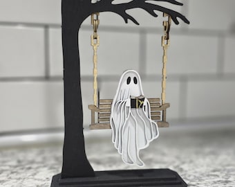 May include: A whimsical Halloween decoration featuring a layered wooden ghost on a swing. The ghost is reading a book and suspended from a black tree-shaped frame with gold-colored swing chains. The base is black, and the ghost is white.
