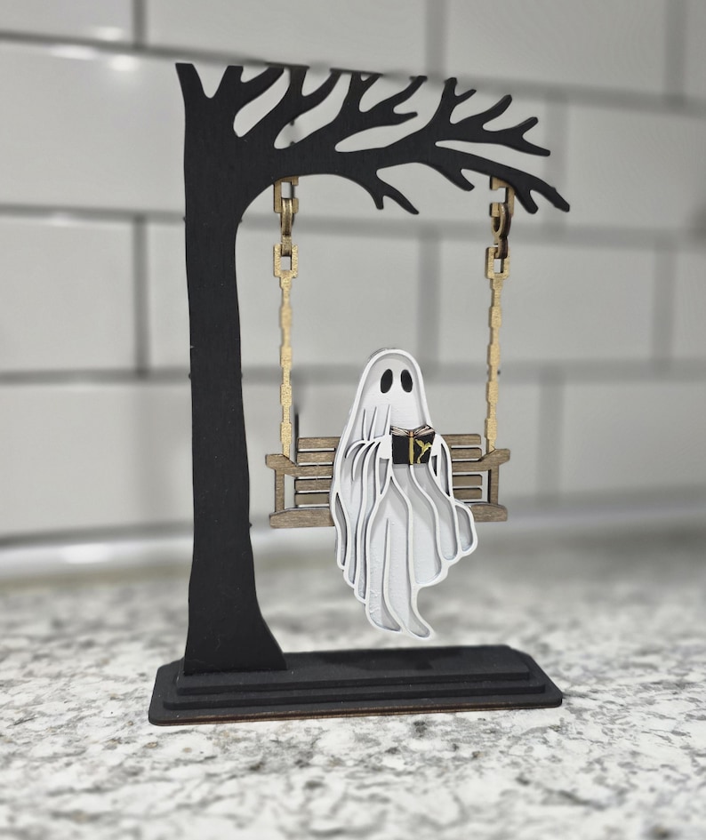May include: A whimsical Halloween decoration featuring a layered wooden ghost on a swing. The ghost is reading a book and suspended from a black tree-shaped frame with gold-colored swing chains. The base is black, and the ghost is white.