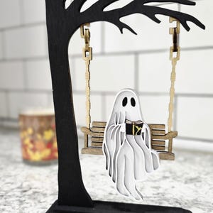 May include: A wooden Halloween decoration featuring a black tree with a swing. A white ghost reading a book sits on the swing. The tree and swing are made of wood and have a layered design.