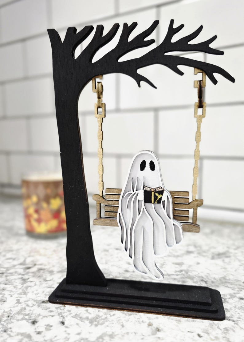 May include: A wooden Halloween decoration featuring a black tree with a swing. A white ghost reading a book sits on the swing. The tree and swing are made of wood and have a layered design.