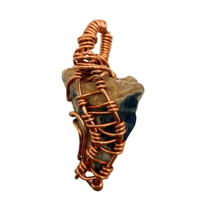 Puede incluir: Colgante artesanal con una piedra natural en tonos azules y beige, envuelta en alambre de cobre. El alambre forma una jaula decorativa alrededor de la piedra, con un lazo para una cadena. El colgante se muestra sobre un fondo blanco.