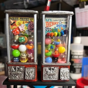 May include: Two vintage-style gumball machines, one red with a "Magic Tricks & Pranks" sign and the other red with a "Lil' Derby Ducks" sign. Both machines have silver coin slots and are filled with colorful gumballs.