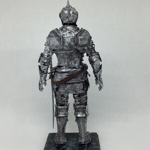 May include: A silver-painted figurine of a knight in full armor, standing on a black base. The knight is holding a sword in its right hand. A penny is in the foreground for scale.