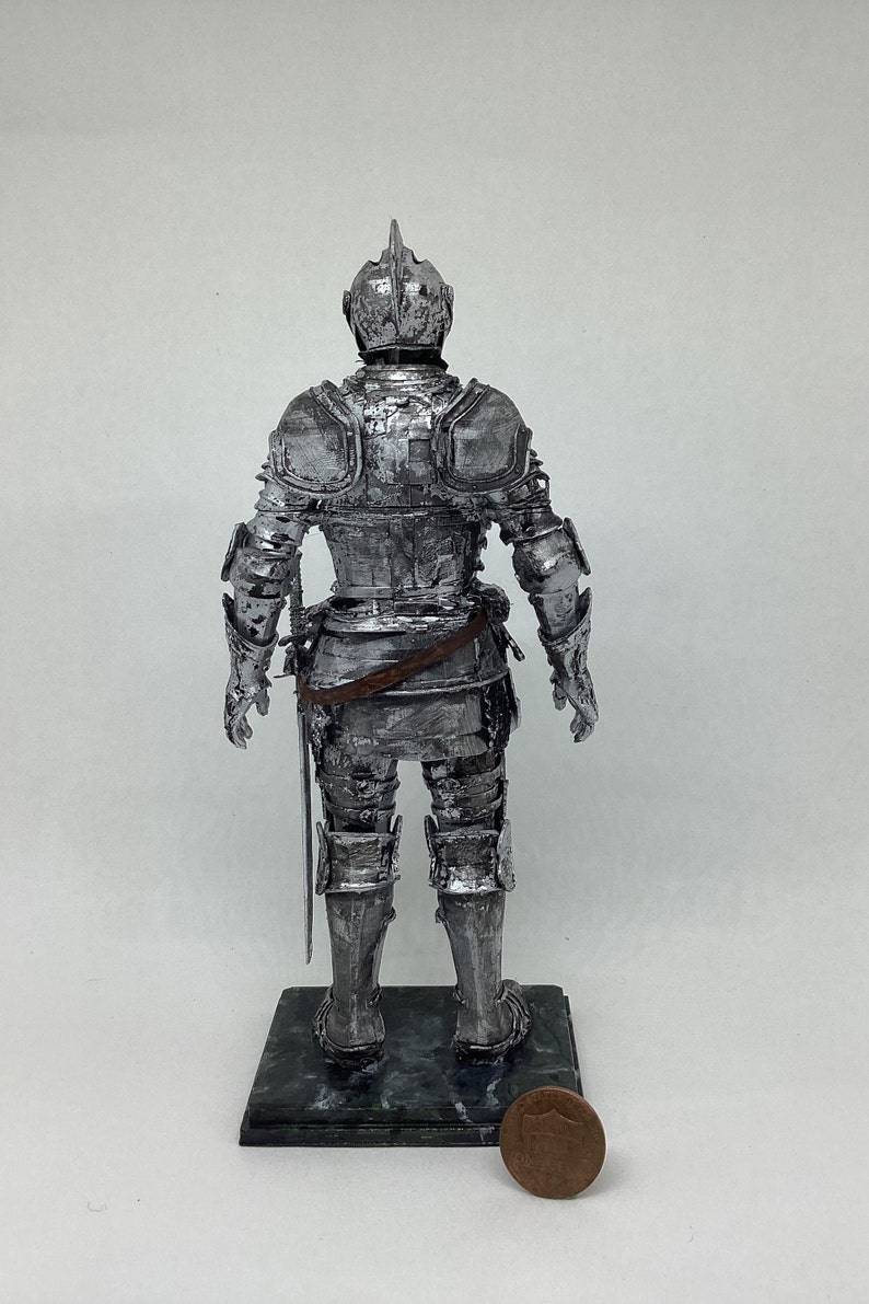 May include: A silver-painted figurine of a knight in full armor, standing on a black base. The knight is holding a sword in its right hand. A penny is in the foreground for scale.