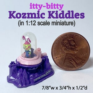 May include: A miniature Kozmic Kiddles figure, 7/8" wide, 3/4" high, and 1/2" deep, featuring a small alien inside a clear dome. The figure is set on a purple base with pink accents. The text "itty-bitty Kozmic Kiddles" is visible.