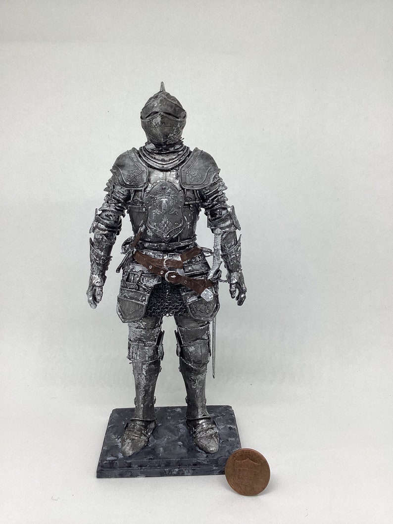 May include: A silver-colored knight figurine wearing armor and holding a sword. The figurine is standing on a black base and is about 4 inches tall.