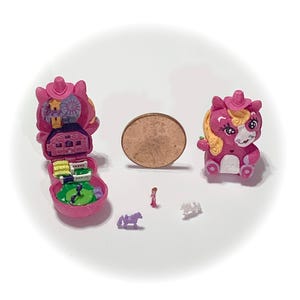 May include: A collection of miniature pink toys, including a horse-shaped figure, a miniature playset, and a pony figure. A penny is included for size comparison. The toys are brightly colored and appear to be made of plastic.