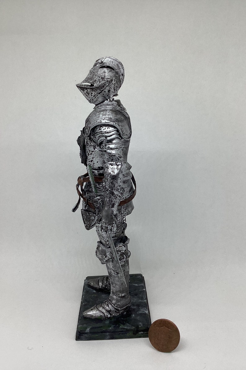 May include: A silver knight figurine standing on a green marble base. The knight is wearing full armor and holding a sword. A penny is in the foreground for scale.