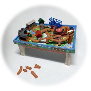 Puede incluir: Un juego de trenes de juguete en miniatura sobre una mesa pintada de azul y blanco. El juego incluye una vía de madera marrón, pequeños edificios, un puente azul y trenes de juguete. La mesa tiene patas blancas y un borde decorativo.