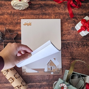Peut inclure: Un bloc-notes avec une illustration de maison en pain d'épices est ouvert, révélant des pages vierges. L'image est entourée de décorations de Noël, dont un rouleau à pâtisserie, une figurine de gnome et une boîte cadeau avec un nœud rouge.