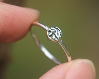 May include: A silver ring with a small, round design featuring a cluster of leaves and acorns.