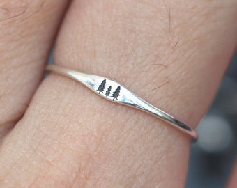 May include: A silver ring with a delicate design of three stylized trees. The ring is worn on a person's finger.