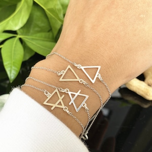 Könnte beinhalten: Drei silberne Armbänder mit Dreiecksanhängern. Die Anhänger sind in verschiedenen Formen und Größen. Die Armbänder sind übereinander am Handgelenk einer Person gelegt.