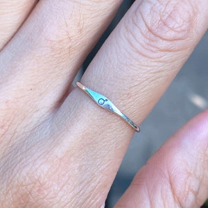 May include: A slender silver ring with a delicate band and an elongated central design. The design features a small, engraved symbol of the planet Mars. The ring is shown on a finger, showcasing its minimalist and elegant style.