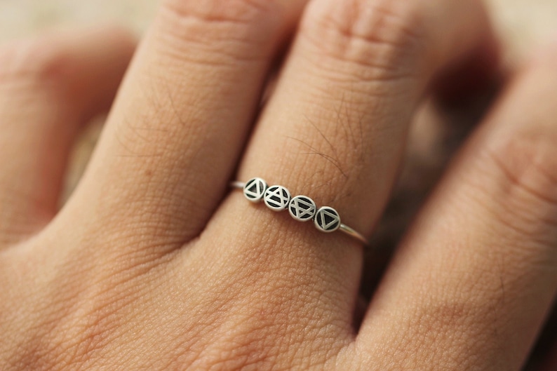 May include: A silver ring with five small charms, each featuring a different geometric design. The charms are connected by a thin silver band.