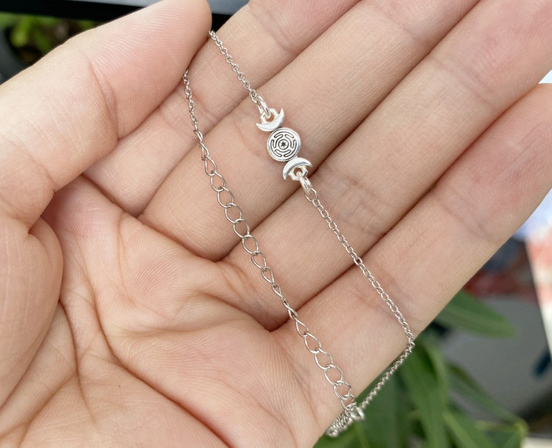 May include: A silver chain necklace with a triple moon pendant. The pendant features a circle with a spiral design inside and three crescent moons.