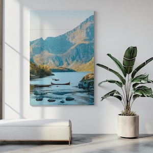 May include: A landscape canvas print featuring a serene lake scene with mountains in the background. Several small boats are on the water. The color palette includes blues, greens, and browns, creating a calming and natural aesthetic. The artwork is displayed on a white wall.