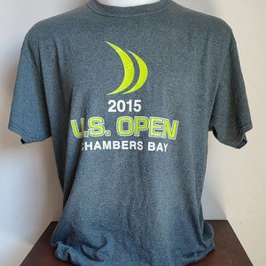 Op de afbeelding: Een donkergrijs T-shirt met een groen logo van drie gebogen lijnen en de tekst "2015 U.S. OPEN CHAMBERS BAY".