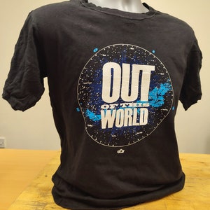 Può includere: Maglietta nera con una grafica blu e bianca di un cielo stellato con il testo "OUT OF THIS WORLD".