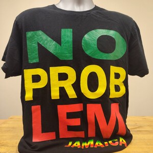 以下が含まれることがあります： 緑、黄色、赤の文字で「NO PROBLEM」と書かれた黒いTシャツ。フレーズの真下に緑、黄色、赤の文字で「JAMAICA」と印刷されています。