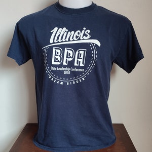 Puede incluir: Una camiseta azul marino con un gráfico blanco de un círculo con el texto "Illinois BPA State Leadership Conference 2018 Dream Bigger" en su interior.