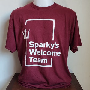 Könnte beinhalten: Ein maroonfarbenes T-Shirt mit einer weißen Grafik eines Quadrats mit einem Dreizack und dem Text "Sparky's Welcome Team" darin.