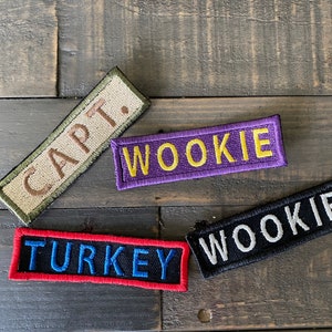 May include: Four embroidered patches with text. One reads "CAPT." in brown on a tan background with a green border. Another reads "WOOKIE" in yellow on a purple background. A third reads "TURKEY" in blue on a black background with a red border. The last reads "WOOKIE" in white on a black background.