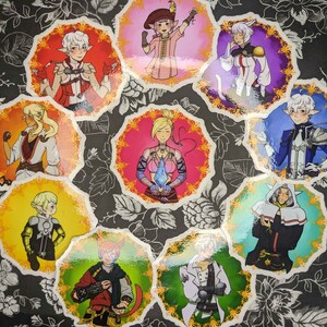 Puede incluir: Nueve pegatinas coloridas con ilustraciones de personajes en varias poses. Cada pegatina es circular y tiene un borde dorado adornado. Los personajes llevan diferentes atuendos y tienen diferentes peinados. Las pegatinas están sobre un fondo floral blanco y negro.