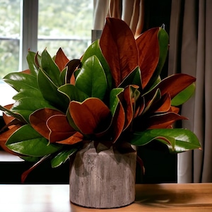 May include: A gray ceramic vase filled with a lush arrangement of green and brown magnolia leaves. The vase has a textured, ribbed surface. The text "Fresh Magnolia Centerpiece" is displayed below the vase.