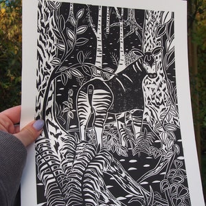 May include: Black and white linocut print of a zebra standing in a forest. The zebra is surrounded by trees and ferns. The print is on white paper.