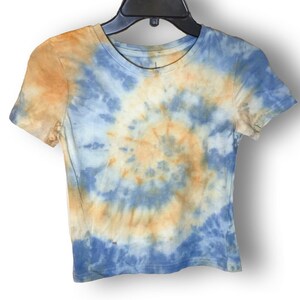 May include: A short-sleeved t-shirt with a tie-dye pattern in shades of blue and orange. The design spirals from the centre, creating a circular effect. The shirt is hanging on a black hanger against a white background.