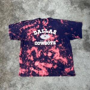 May include: A navy blue t-shirt with a tie-dye effect in pink and white. The shirt features the text "DALLAS HOME OF THE COWBOYS" in white, with a football graphic in the center. The shirt is a classic crew neck style.