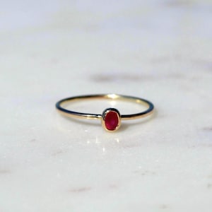 May include: A delicate gold ring featuring a vibrant oval-cut ruby gemstone. The ring's slender band and bezel setting highlight the rich red color of the gemstone. The ring is displayed on a white marble surface.