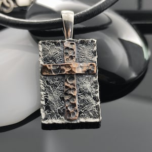 May include: A silver pendant necklace with a rectangular cross pendant. The cross is made of hammered silver and copper, with a textured finish.