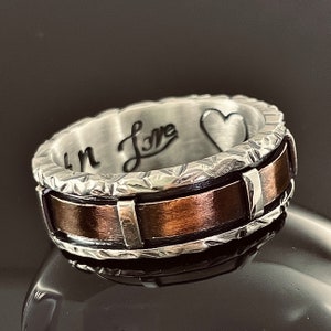 Op de afbeelding: Een zilveren en koperen bandring met een getextureerde rand en een gegraveerd hart aan de binnenkant. De inscriptie "In Love" is ook gegraveerd aan de binnenkant van de ring.