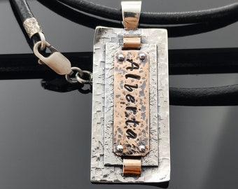 Puede incluir: Collar con colgante de plata y cobre con la palabra "Alberta" grabada en una placa de cobre. El colgante está unido a un cordón de cuero negro.