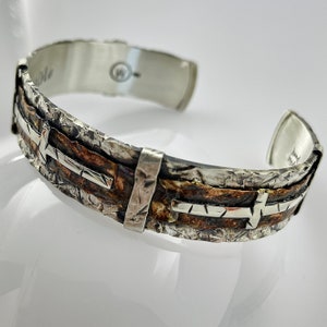 May include: Silver and copper cuff bracelet with a hammered texture and a cross design. The bracelet is open and has a hinged clasp.