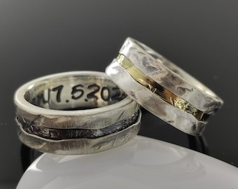 Puede incluir: Dos alianzas de plata con textura martillada. Una alianza tiene una fina banda de oro incrustada en la plata. La otra alianza tiene una banda negra incrustada en la plata. La alianza con la banda de oro tiene la fecha "1.5.2023" grabada en el interior.