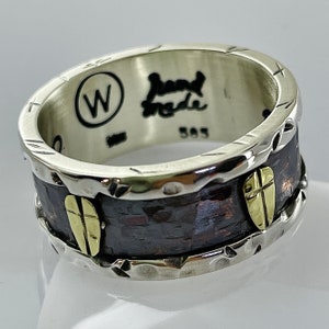 Op de afbeelding: Een zilveren en koperen ring met een gehamerde textuur en een gouden kruisontwerp. De ring is gegraveerd met "Hand Made" en "W".